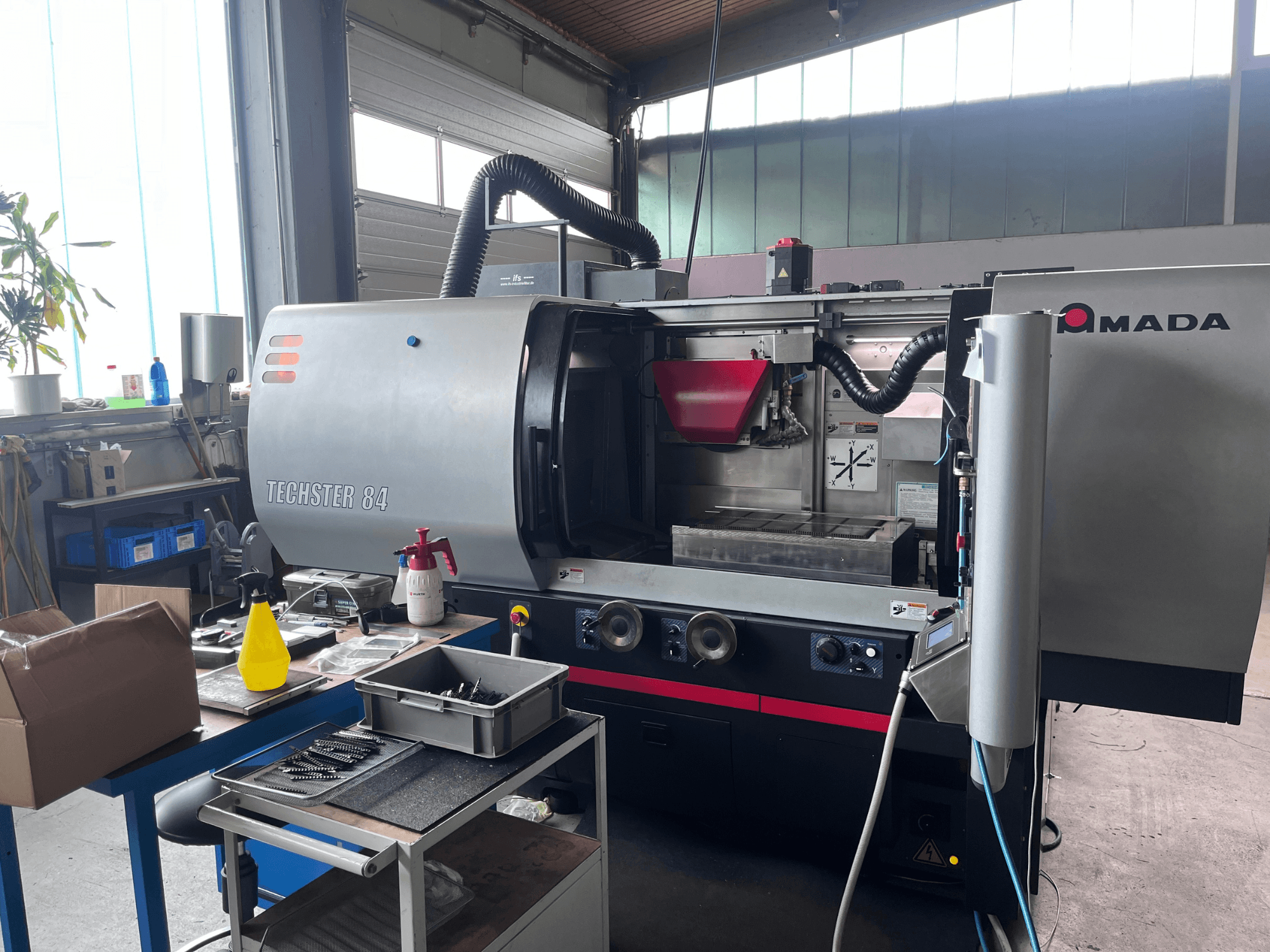 Поверхнево-шліфувальний верстат AMADA Techster 84 у цеху: вид спереду з елементами управління та червоним бункером.