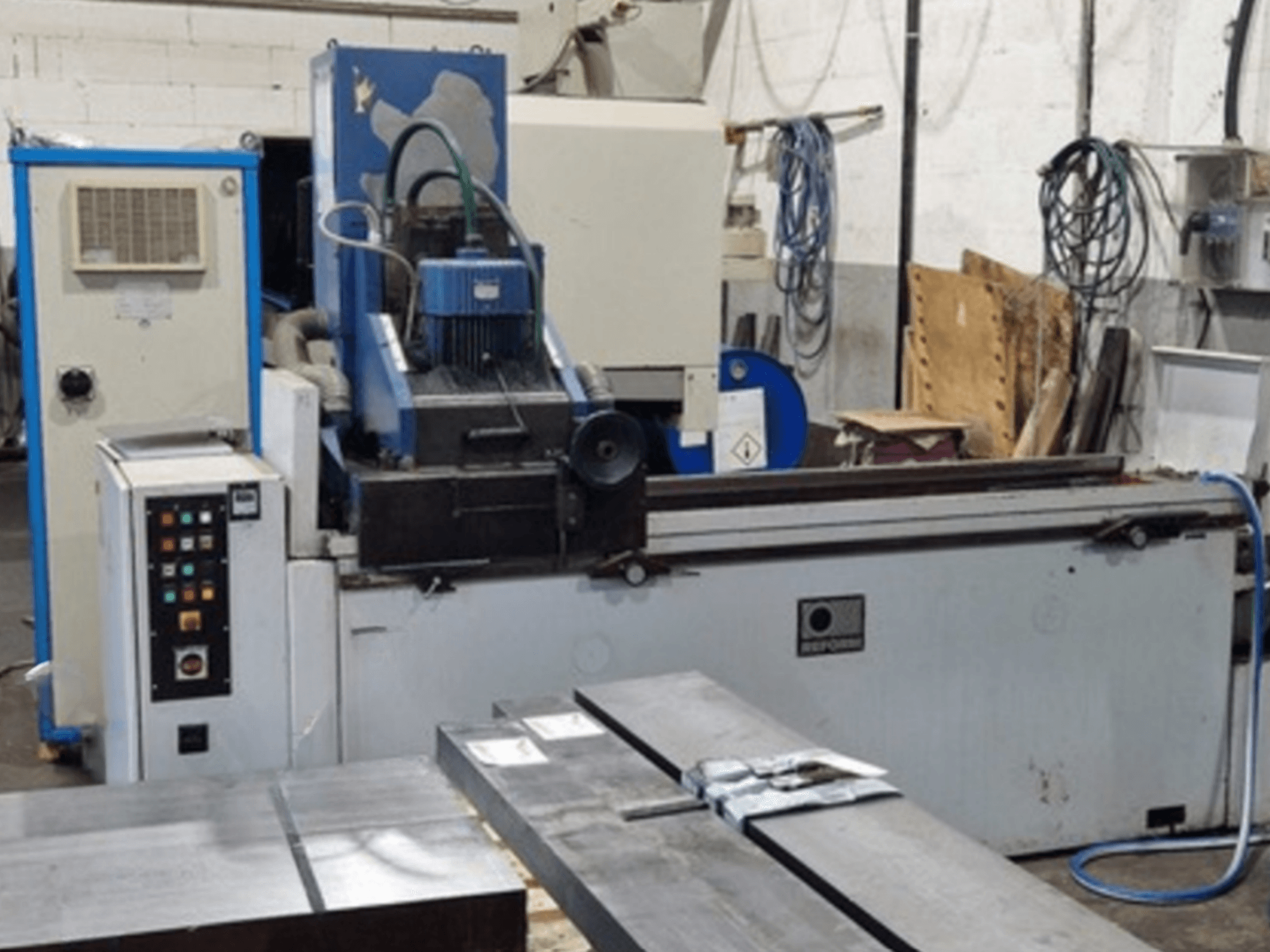 Шліфувальна машина Reform Surface Grinder спереду, з панеллю керування, зоною шліфування та металевими заготовками на столі.