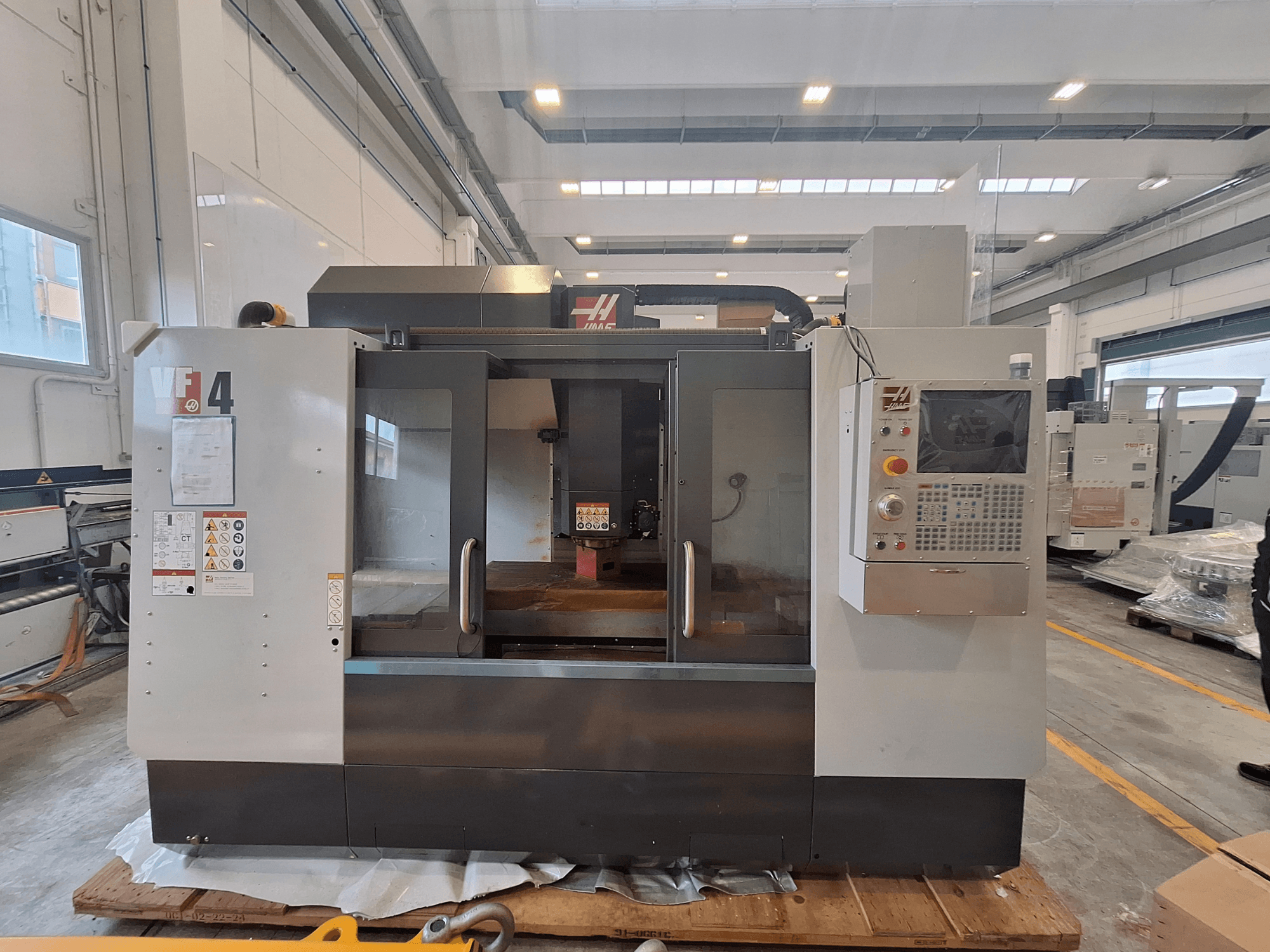Вертикальний обробний центр HAAS VF-4, вид спереду, на якому видно панель управління, робочу зону та попереджувальні написи в умовах складу.