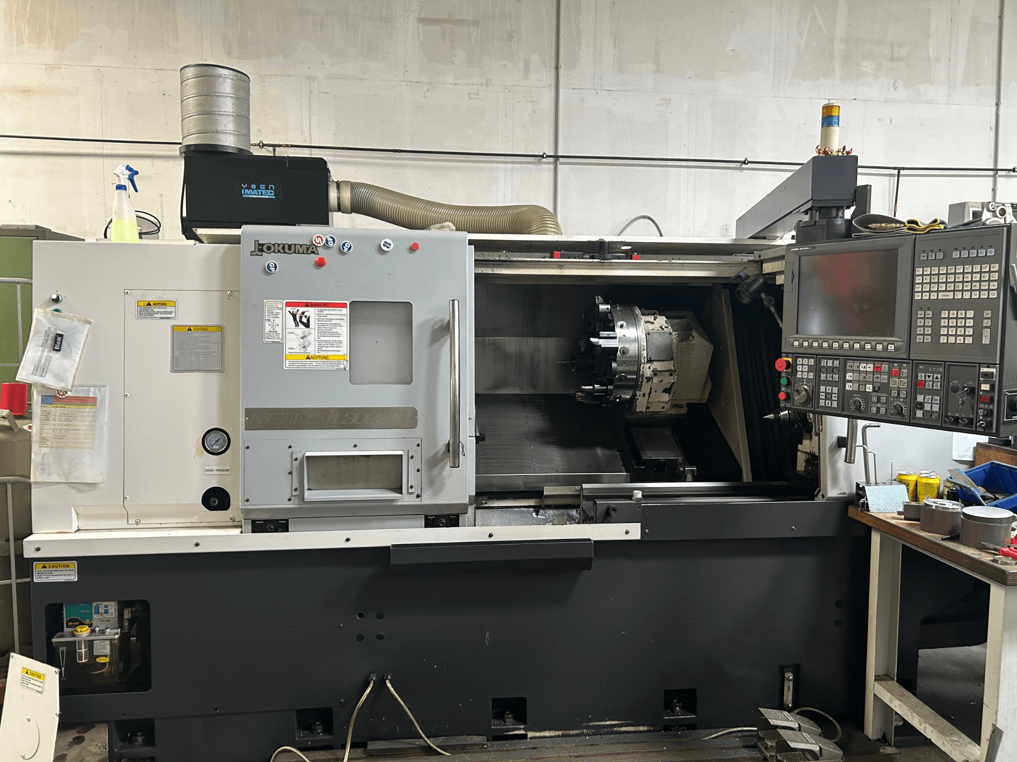Вид спереду горизонтального токарного верстата Okuma Genos L300E-M з панеллю керування, видимим шпинделем і різними інструментами для налаштування.
