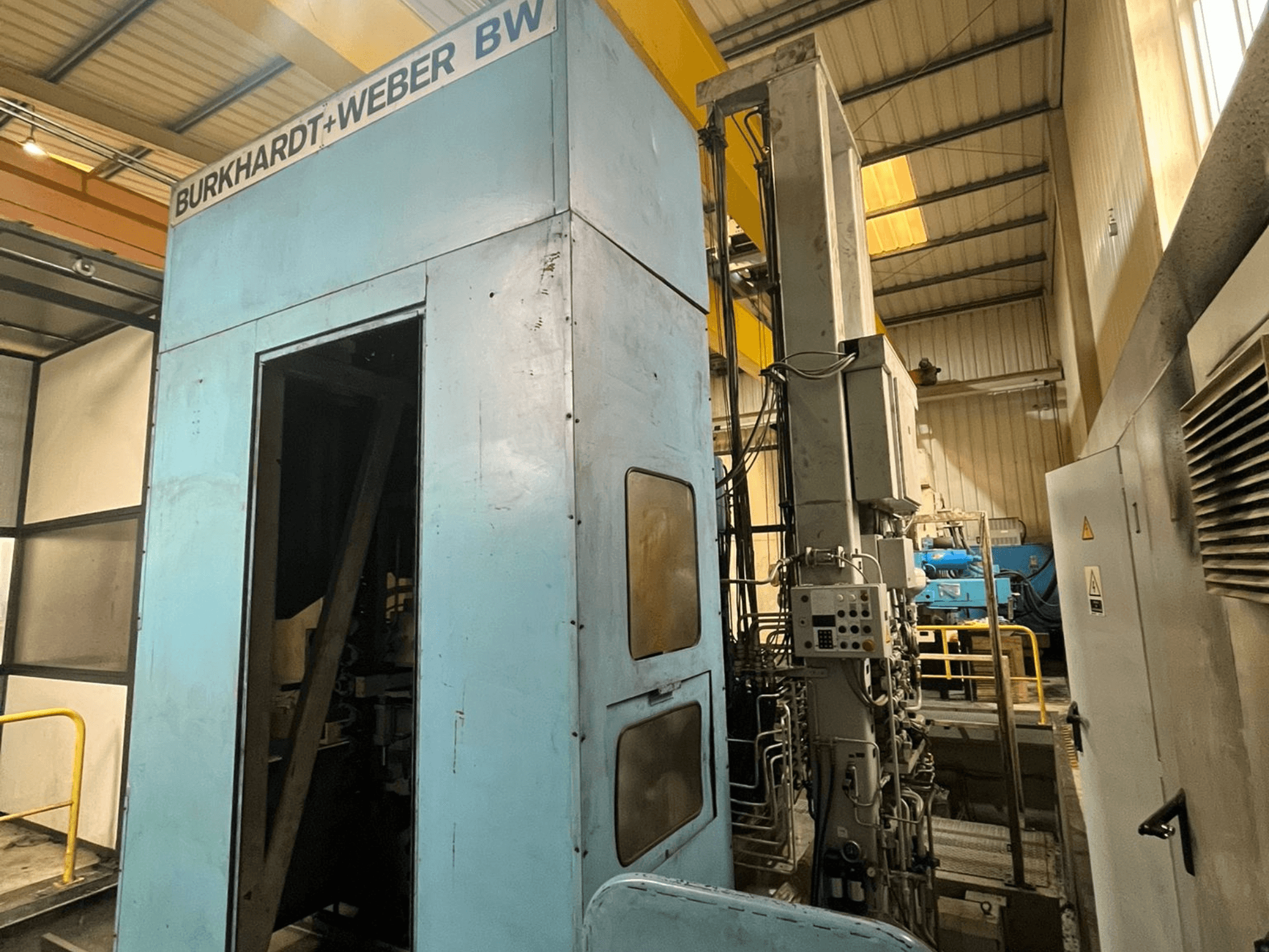 Горизонтальний обробний центр Burkhardt-Weber MC100, вид збоку, на якому видно синій корпус та панель управління з відкритими трубопроводами.
