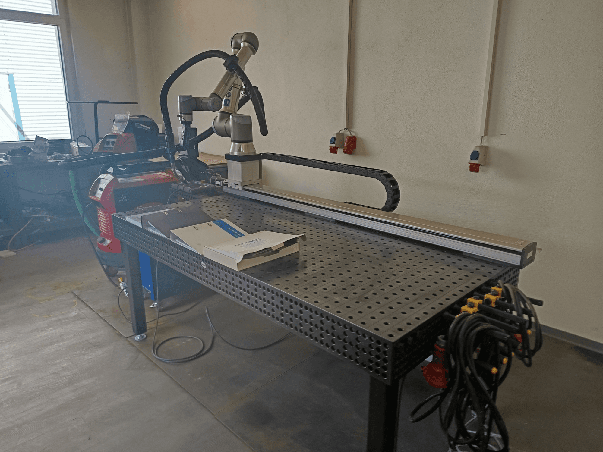 Імпульсна зварювальна установка FRONIUS TransSteel 4000, вид збоку, з робочим маніпулятором, робочою зоною та підключеннями до джерела живлення.