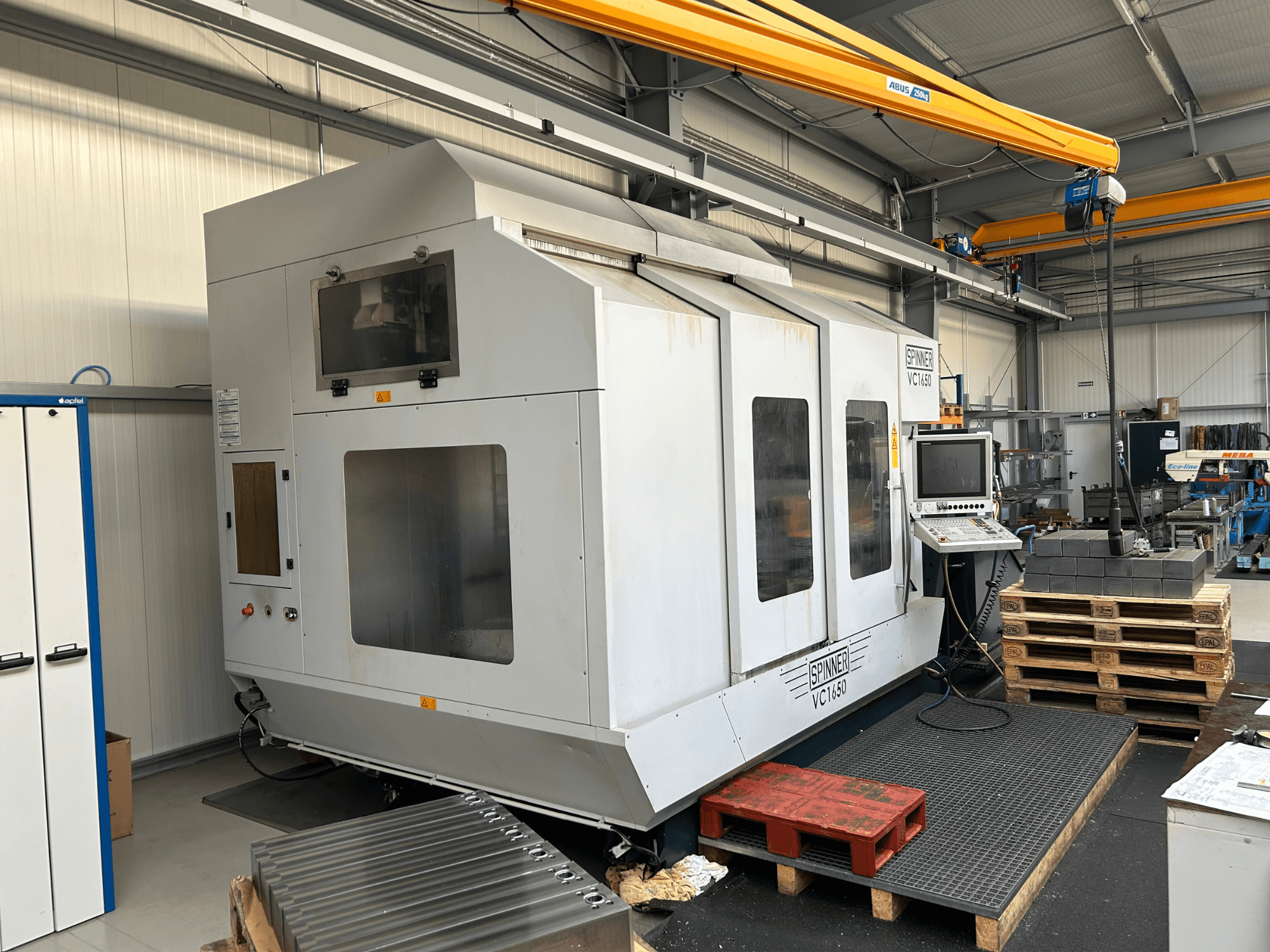 Прядильна машина VC 1650 з ЧПУ, вид спереду, з панеллю керування, великими вікнами та палетами, складеними поруч у цеху.