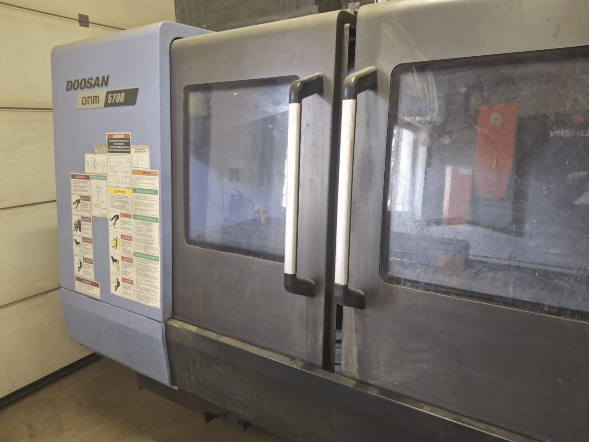Вид спереду вертикального обробного центру DOOSAN DNM 5700 з подвійними дверима та панелями керування з інструкціями з експлуатації.