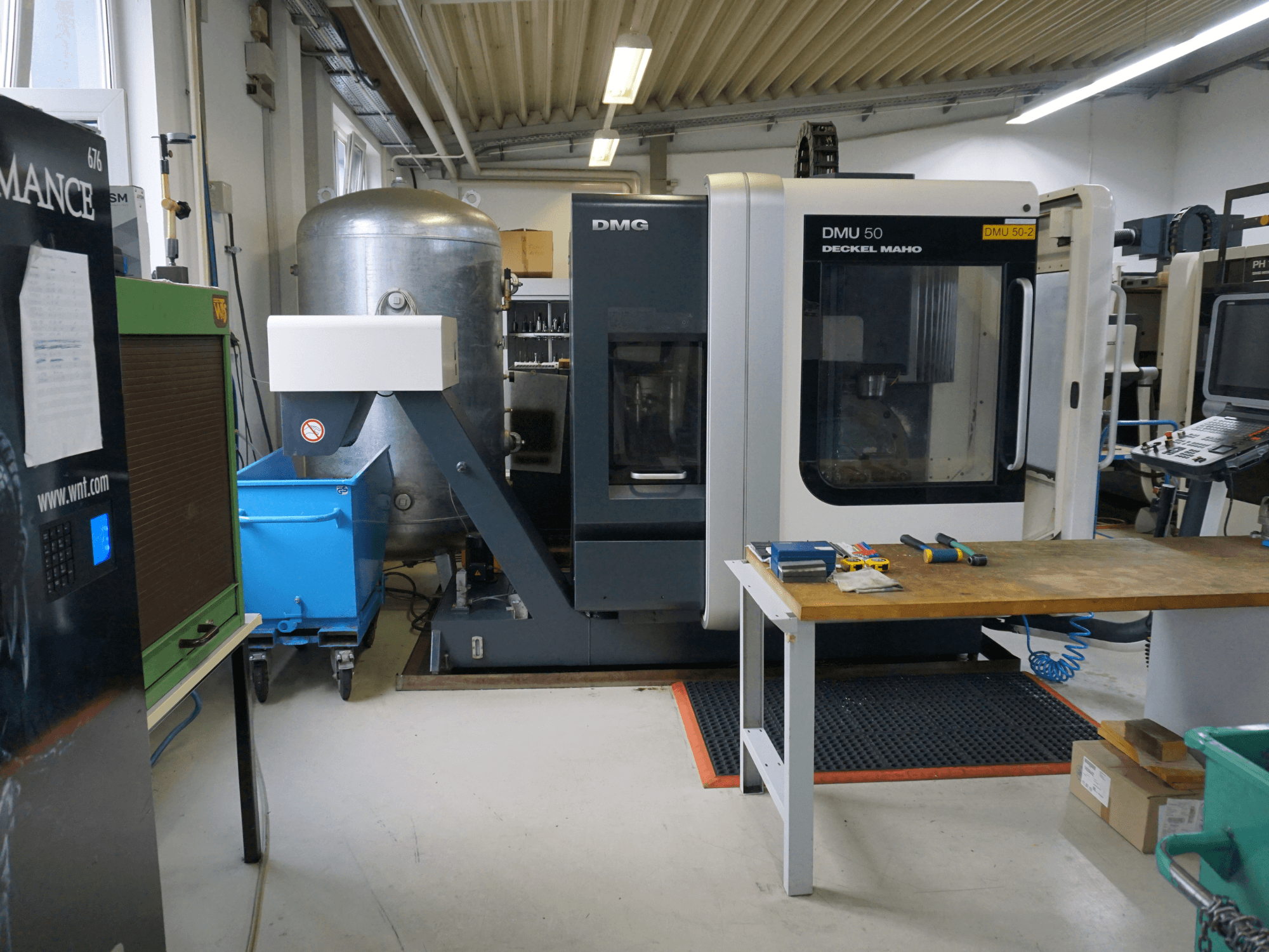 Фрезерний верстат DMG DMU 50 у майстерні, вид спереду, з інструментами на дерев'яному столі та синім контейнером на колесах поруч.