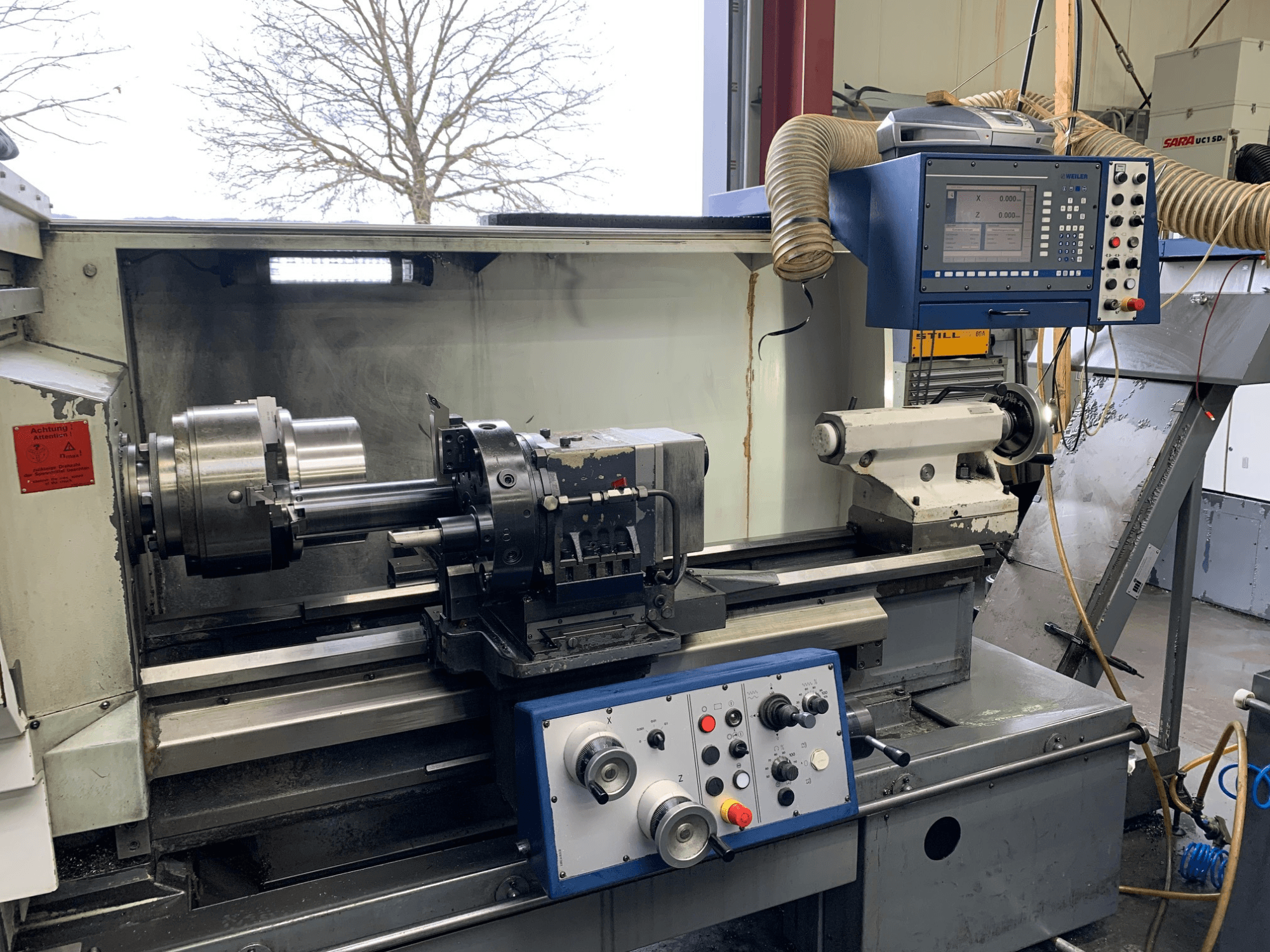 Горизонтальний токарний верстат Weiler E55X1000 в майстерні, з панеллю керування, токарним верстатом і тримачем інструменту. Вид спереду.