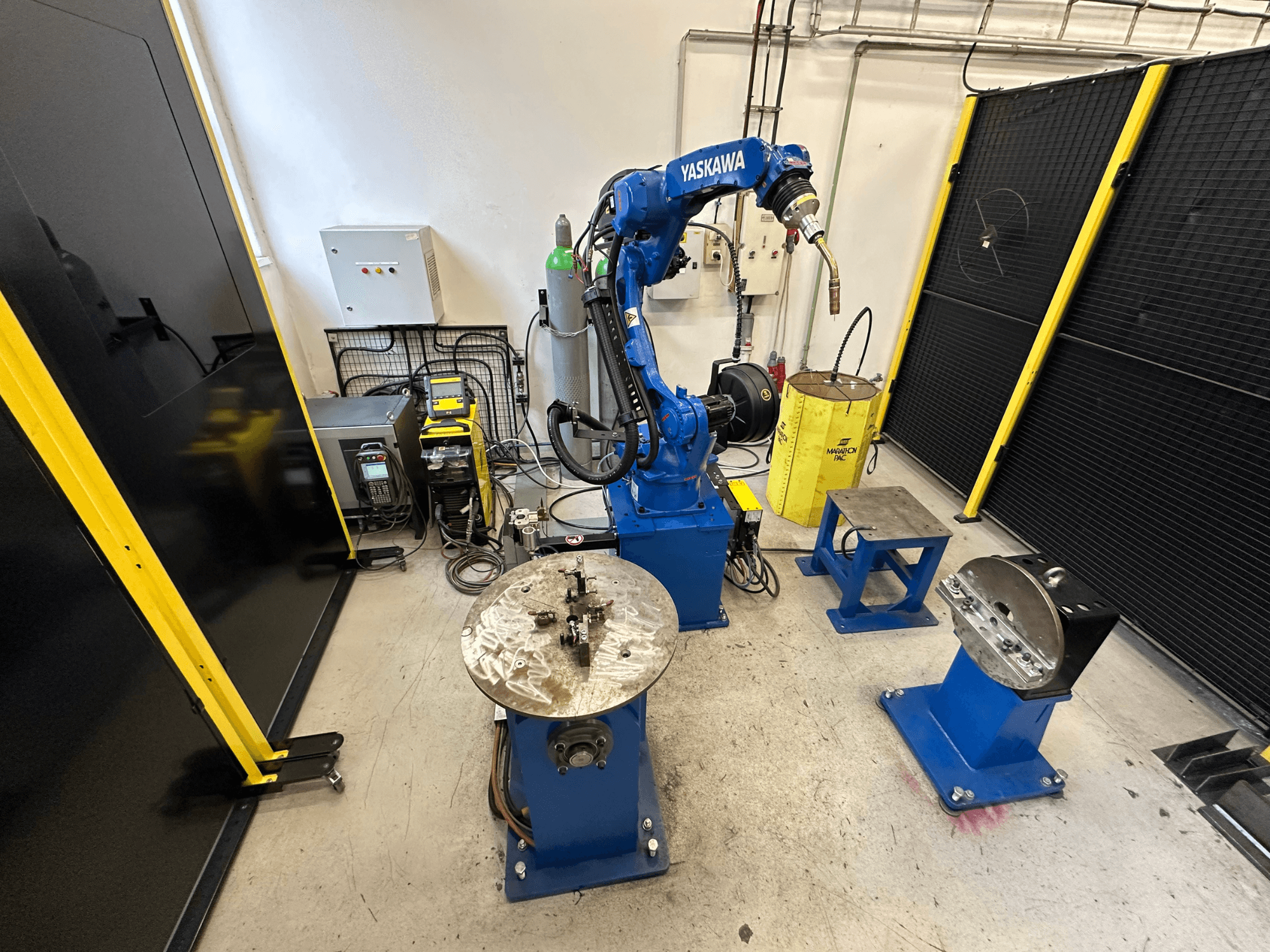 Роботизований маніпулятор Yaskawa синього кольору з двома робочими столами, зварювальним обладнанням та захисними бар'єрами; вид спереду на промисловий робочий простір.