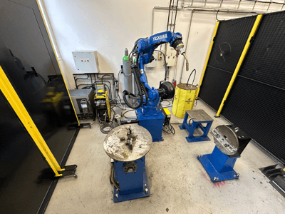 Роботизований маніпулятор Yaskawa синього кольору з двома робочими столами, зварювальним обладнанням та захисними бар'єрами; вид спереду на промисловий робочий простір.