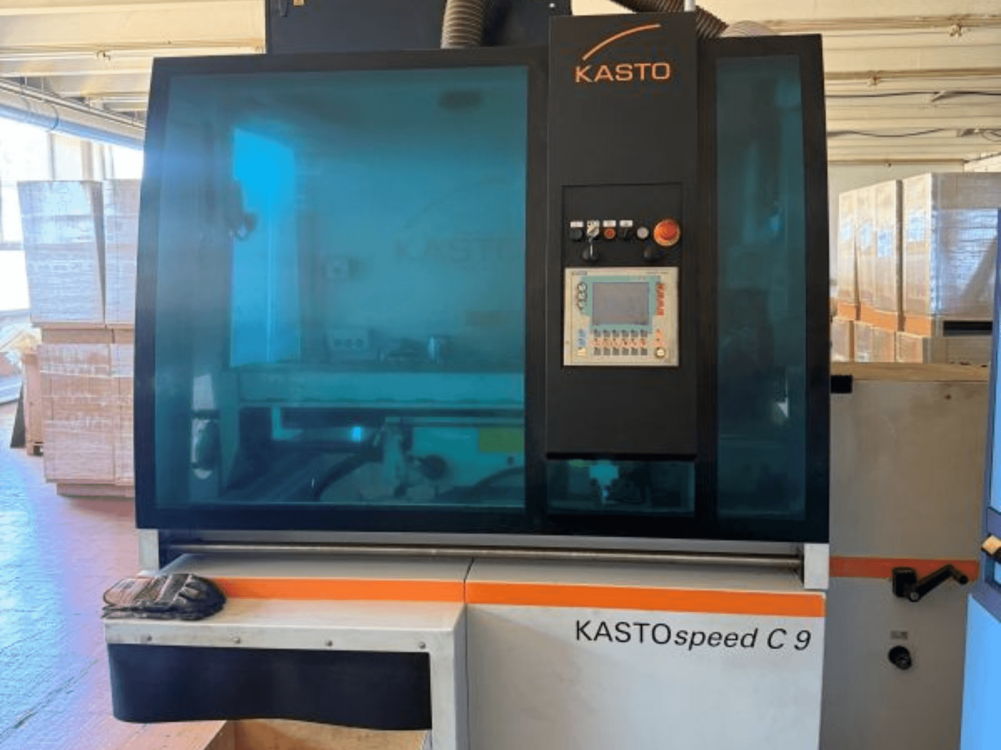 Вигляд спереду циркулярної пилки для металу KASTO KASTOspeed C 9 з панеллю управління та захисним склом; на задньому плані — дерев’яні ящики.