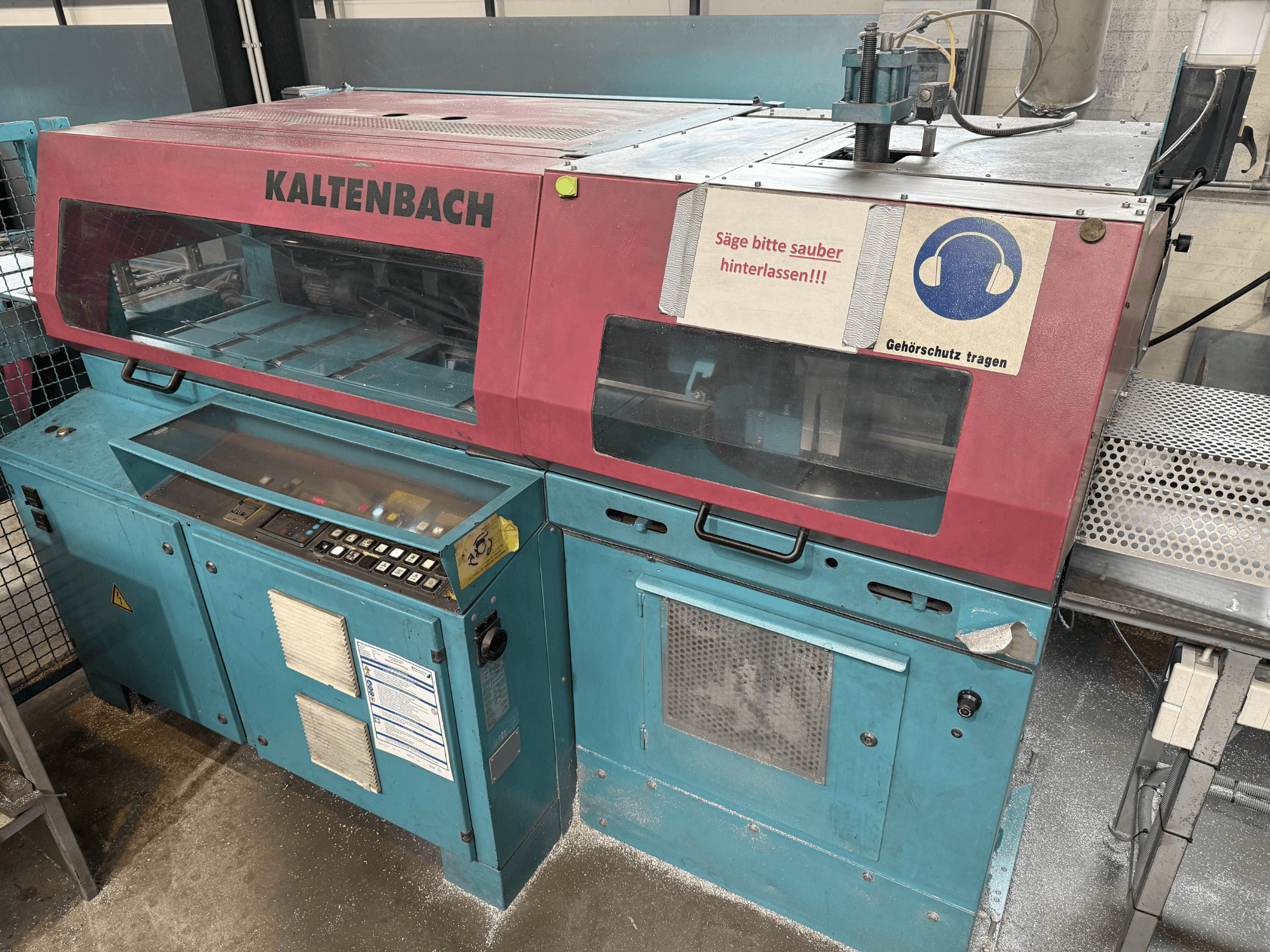 Промисловий розкрійний верстат KALTENBACH, вид спереду, з панеллю керування та знаком безпеки для захисту органів слуху.