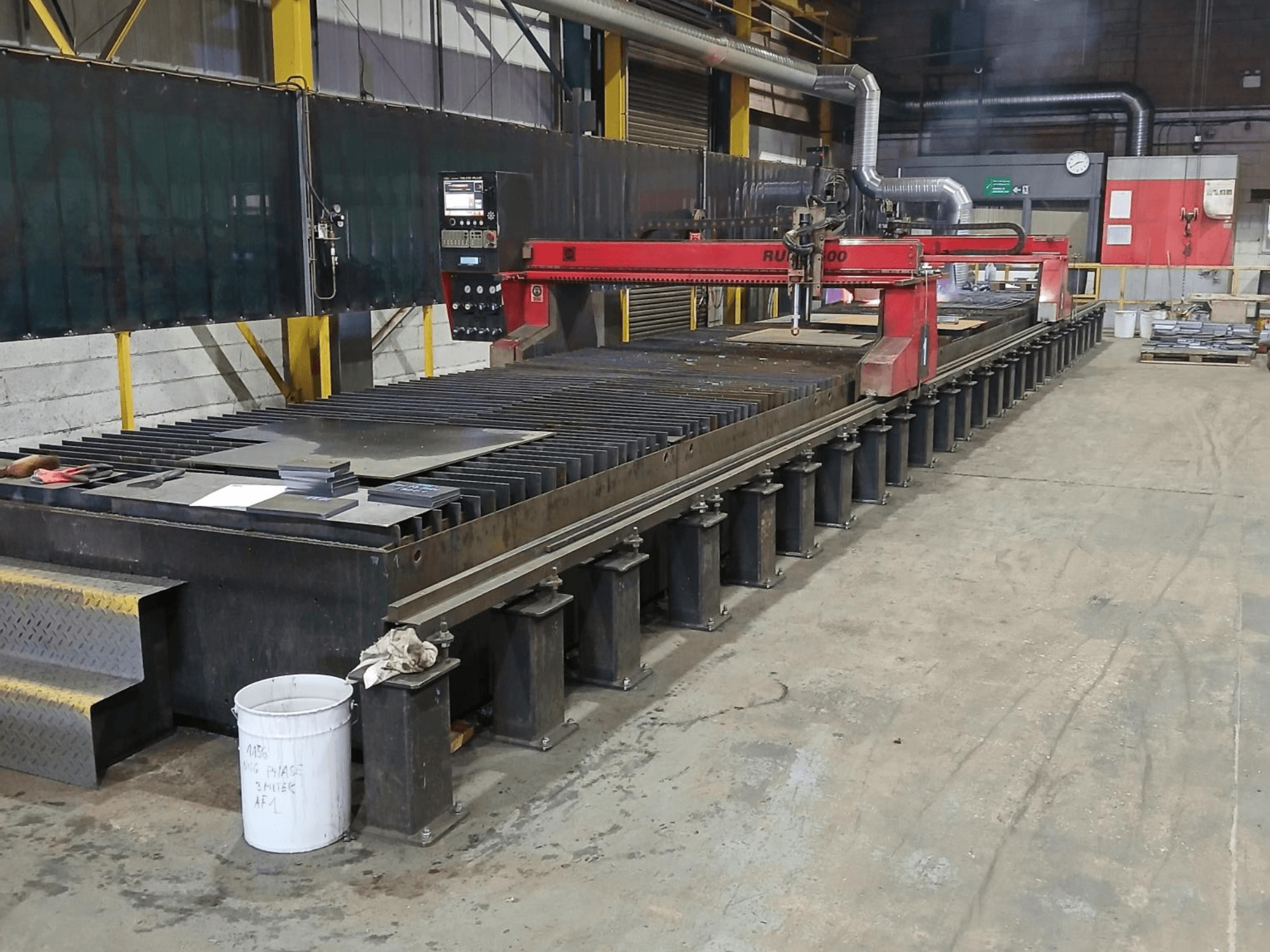 Червоний розкрійний верстат RUIDA 300, вид збоку, з металевим листом на сталевому столі, розташований у промисловому робочому приміщенні.