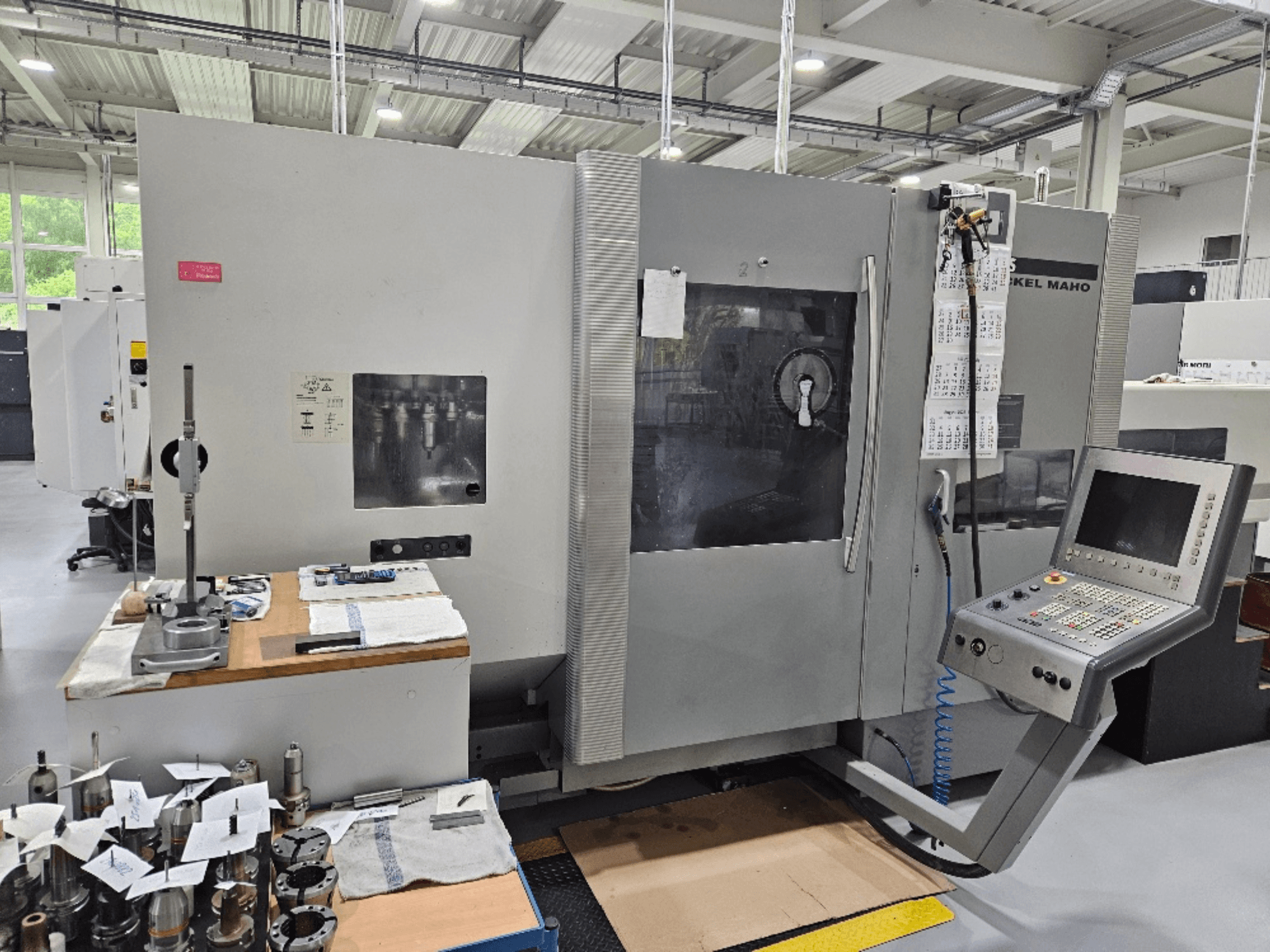 Вид спереду верстата DMG Mori DMU 70 з панеллю керування, інструментами на дерев'яному столі та різними заготовками поруч.