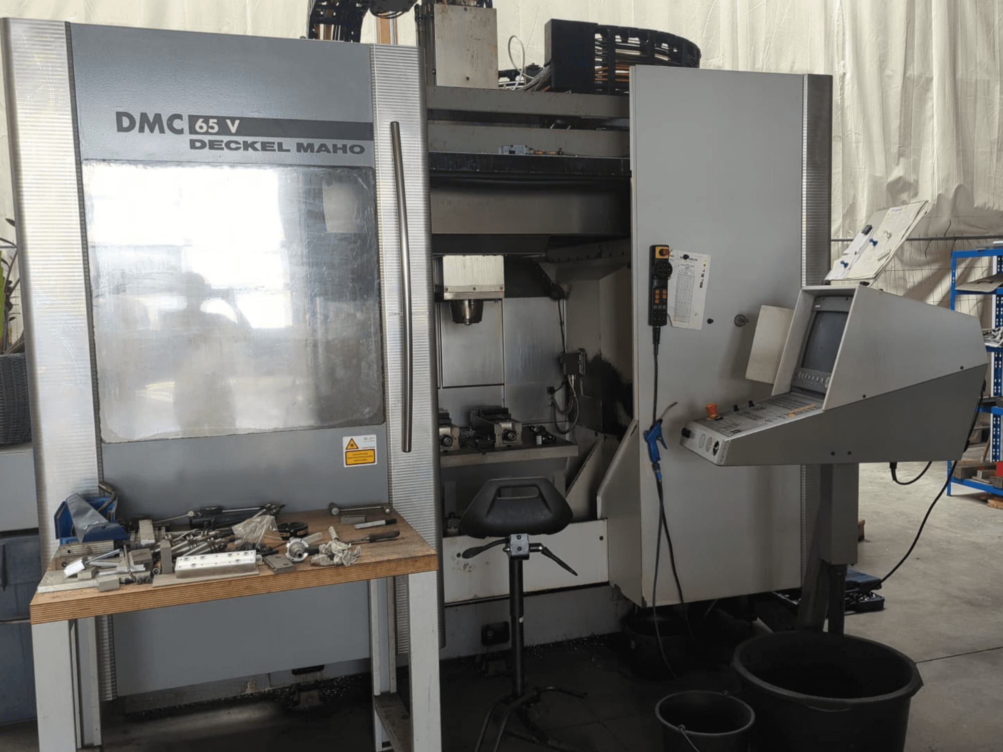 Вигляд спереду вертикального обробного центру DECKEL MAHO DMC 65 V з панеллю керування, інструментами на столі та налаштованим робочим простором.