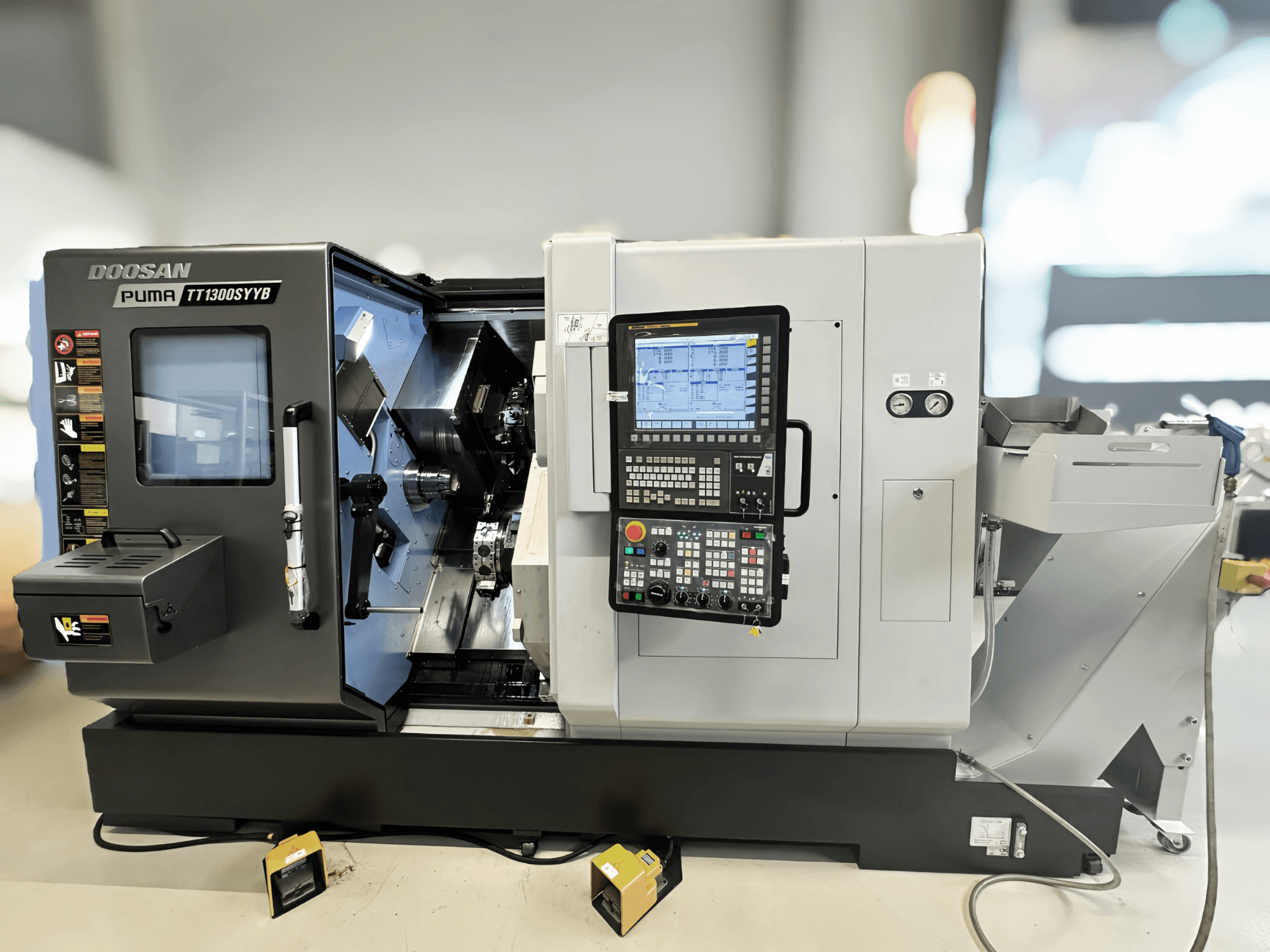 Токарно-фрезерний центр Doosan Puma TT1300SYYB у вигляді збоку, демонструючи панель керування та внутрішню частину верстата з інструментами.