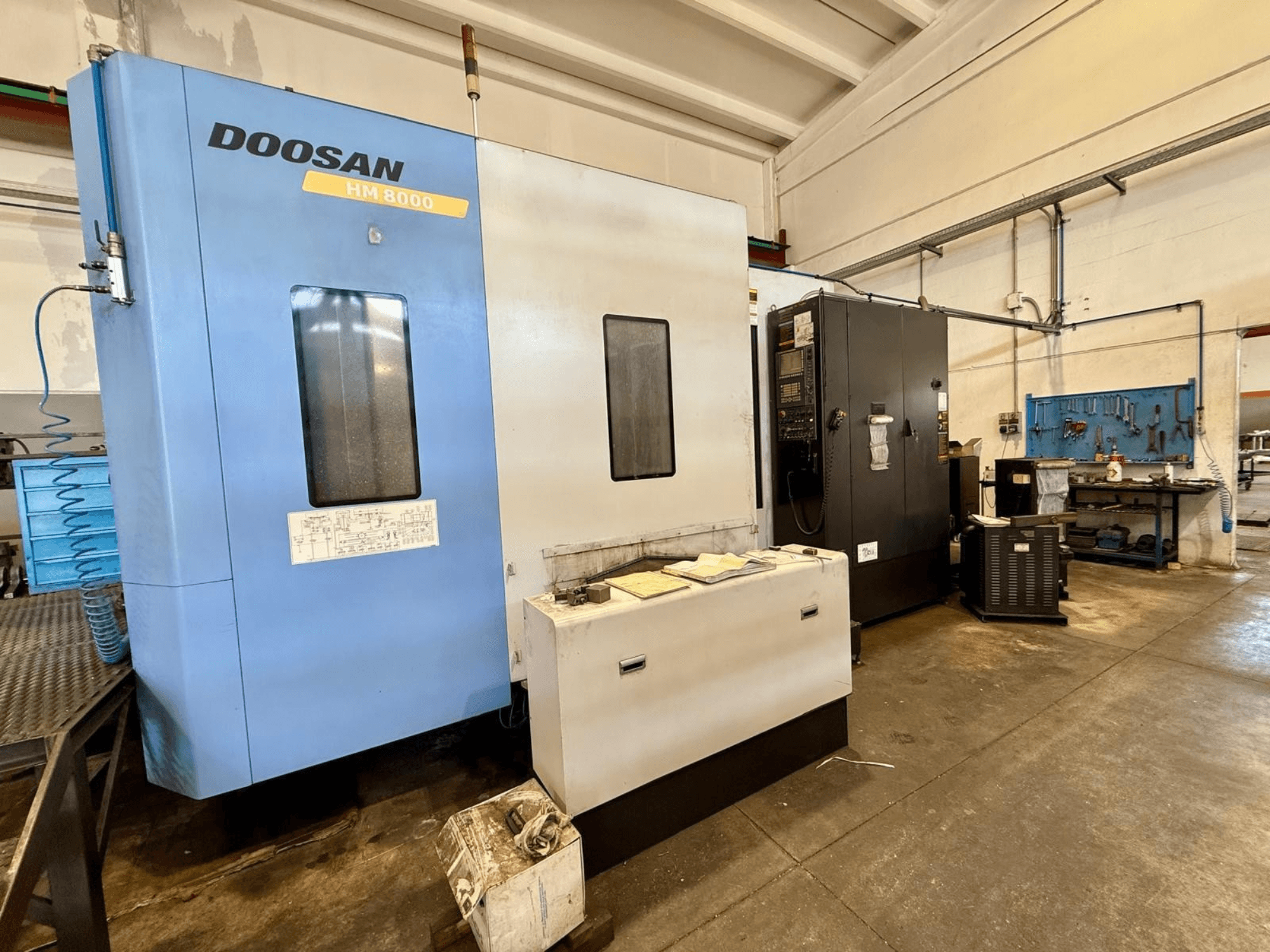 Горизонтальний обробний центр DOOSAN HM 8000 синього кольору, вид спереду, на якому видно панель управління та сусіднє цехове обладнання.