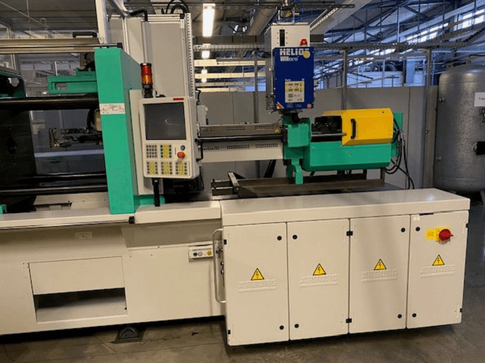 Промислова машина для лиття під тиском, модель HELIO, вигляд спереду, з панеллю керування, захисними кожухами та кнопкою аварійної зупинки.