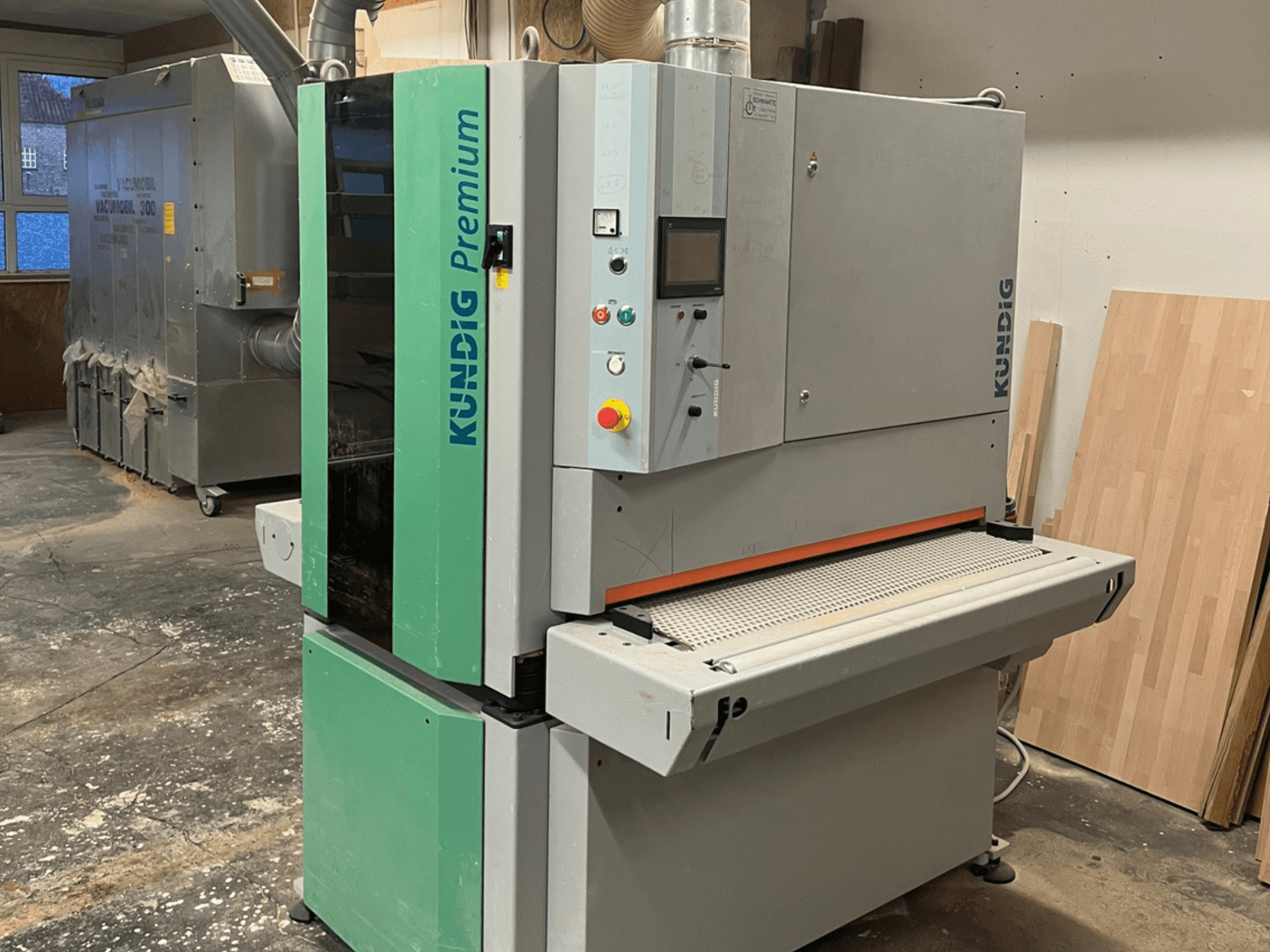 Стрічкова шліфувальна машина Kundig Premium-1 1100K у сірому та зеленому кольорах, показана збоку з панеллю керування та шліфувальною стрічкою.