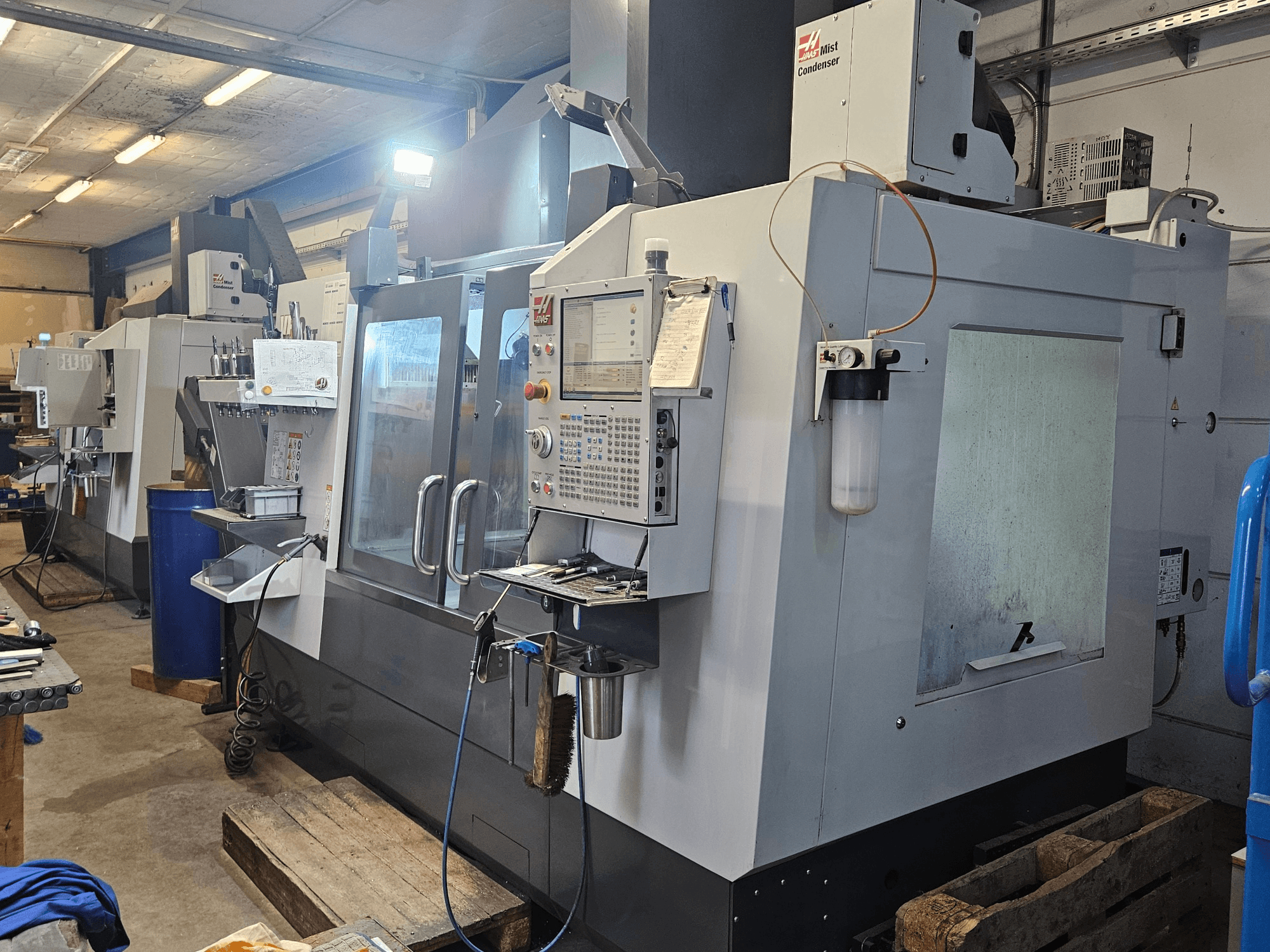 Вертикальний обробний центр Haas VF-3YT/50 у цеху, вид спереду, на якому видно панель управління, тримачі інструментів та конденсатор туману.