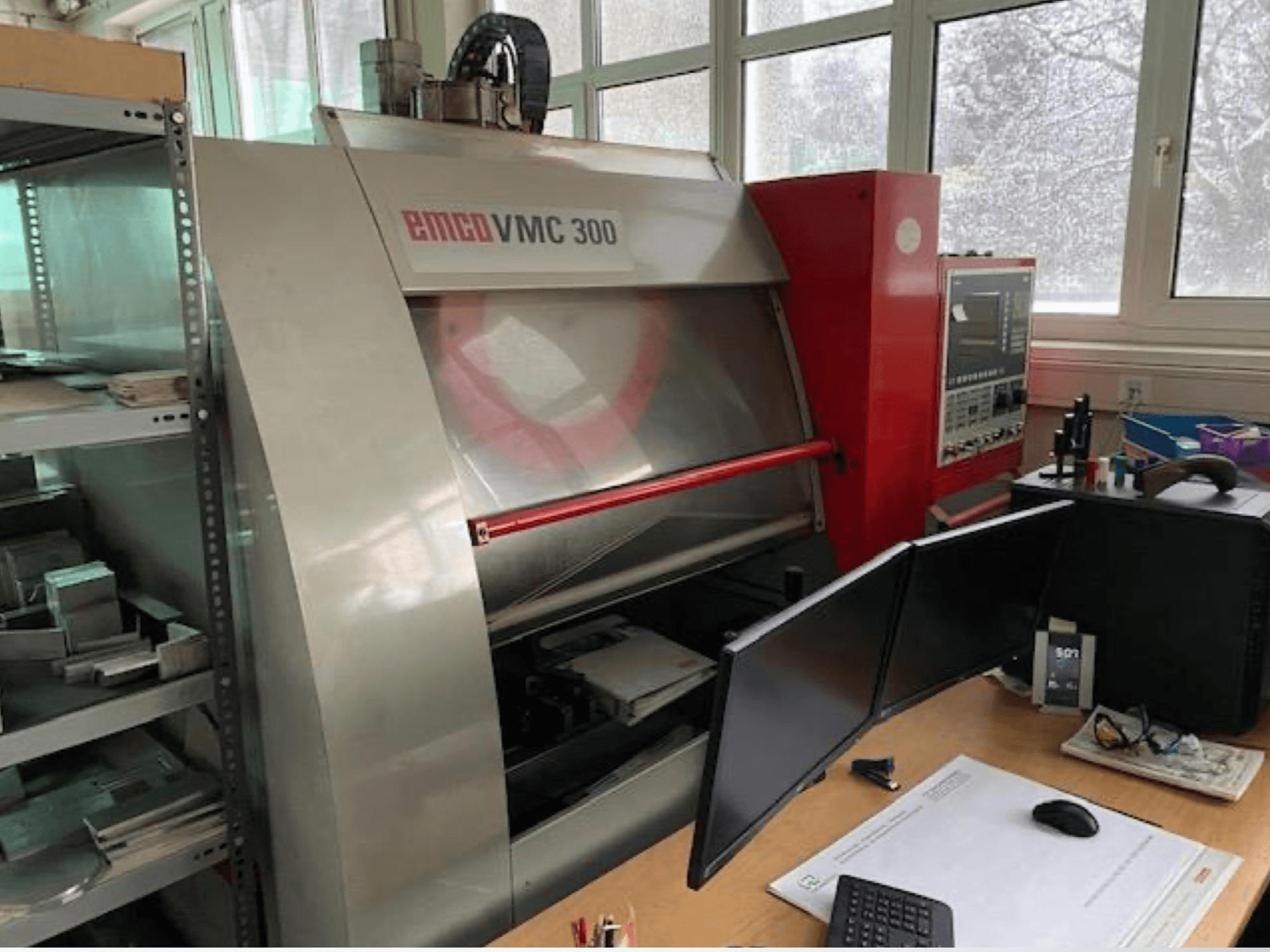 Вертикальний обробний центр EMCO VMC 300 у цеху, вид збоку, на якому видно панель управління та розташоване поруч робоче місце з двома моніторами.