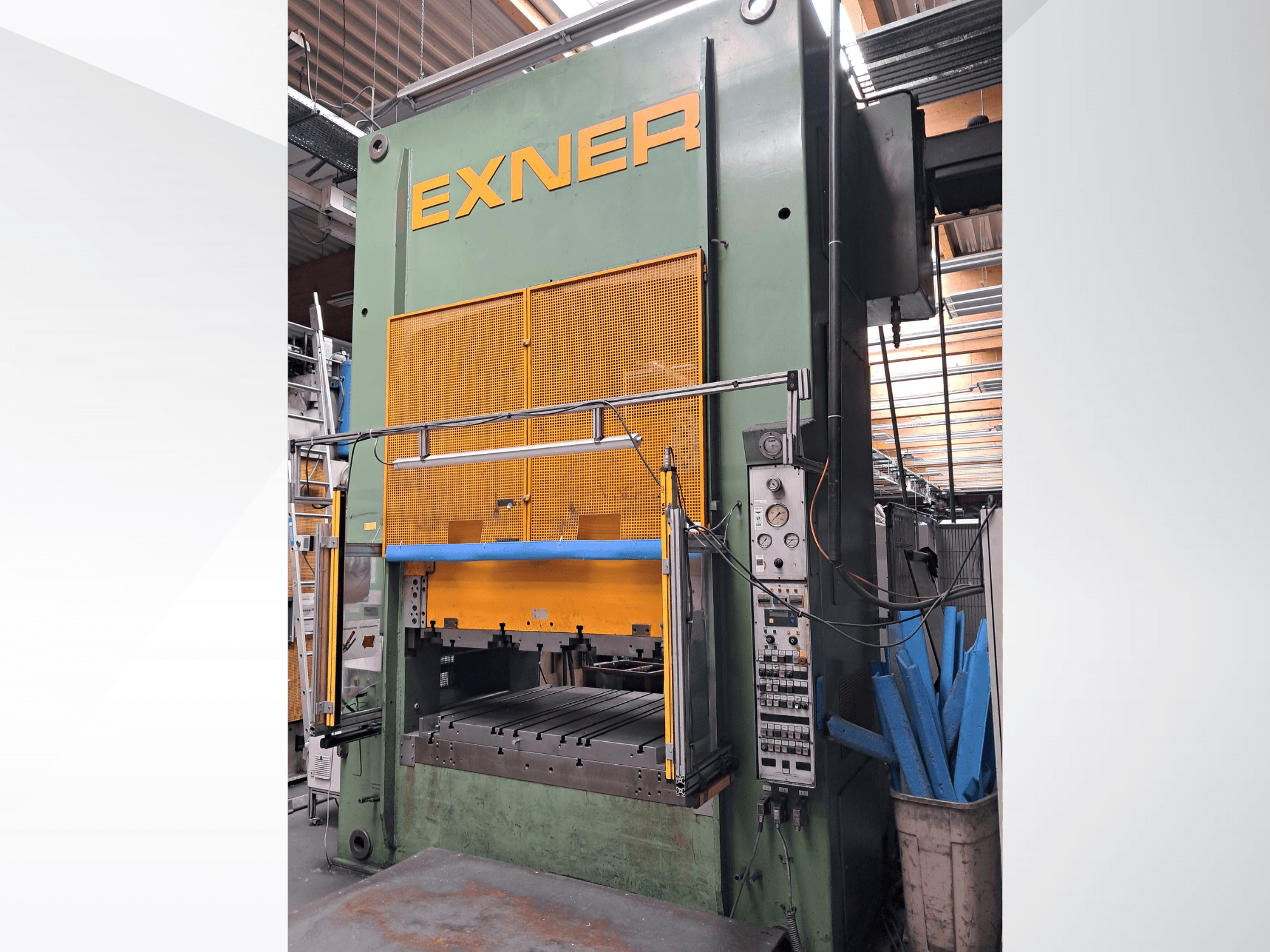 Гідравлічний прес EXNER EX SBR 150, вид спереду; на фото зображено органи управління, синю захисну перекладину та сітчастий захисний кожух; прес встановлено в майстерні.