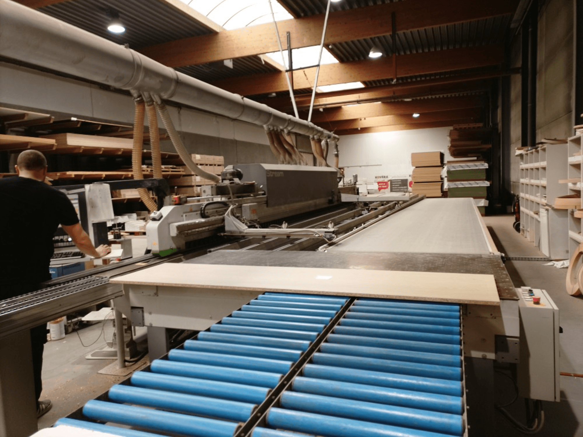 Кромкооблицювальний верстат BIESSE Edge Stream B1, вид збоку: оператор, сині ролики та дерев'яна панель на конвеєрі.