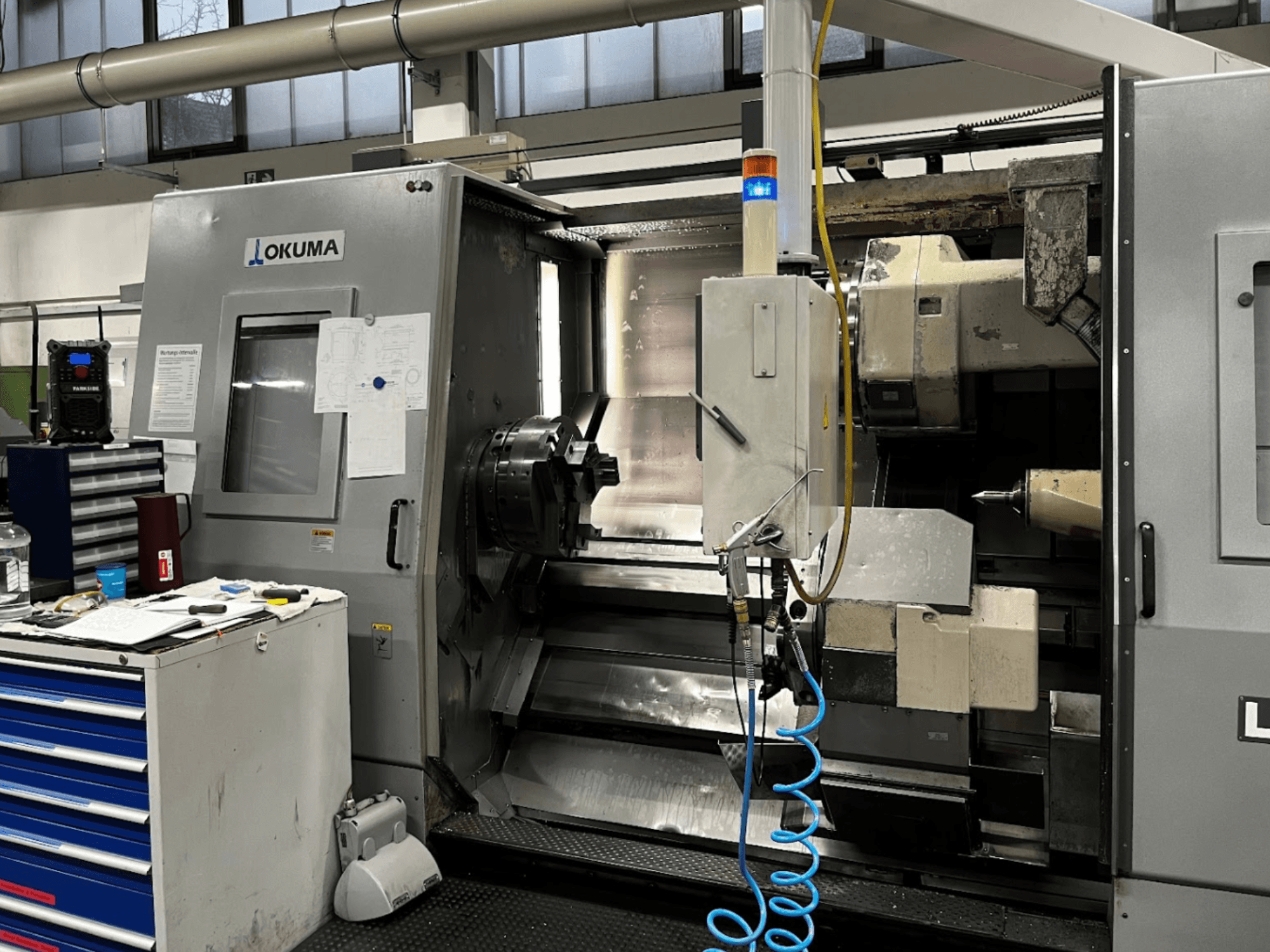 Горизонтальний токарний верстат Okuma LU35-M, вигляд спереду. Оснащений панеллю керування, інструментами та великим патроном для механічної обробки.